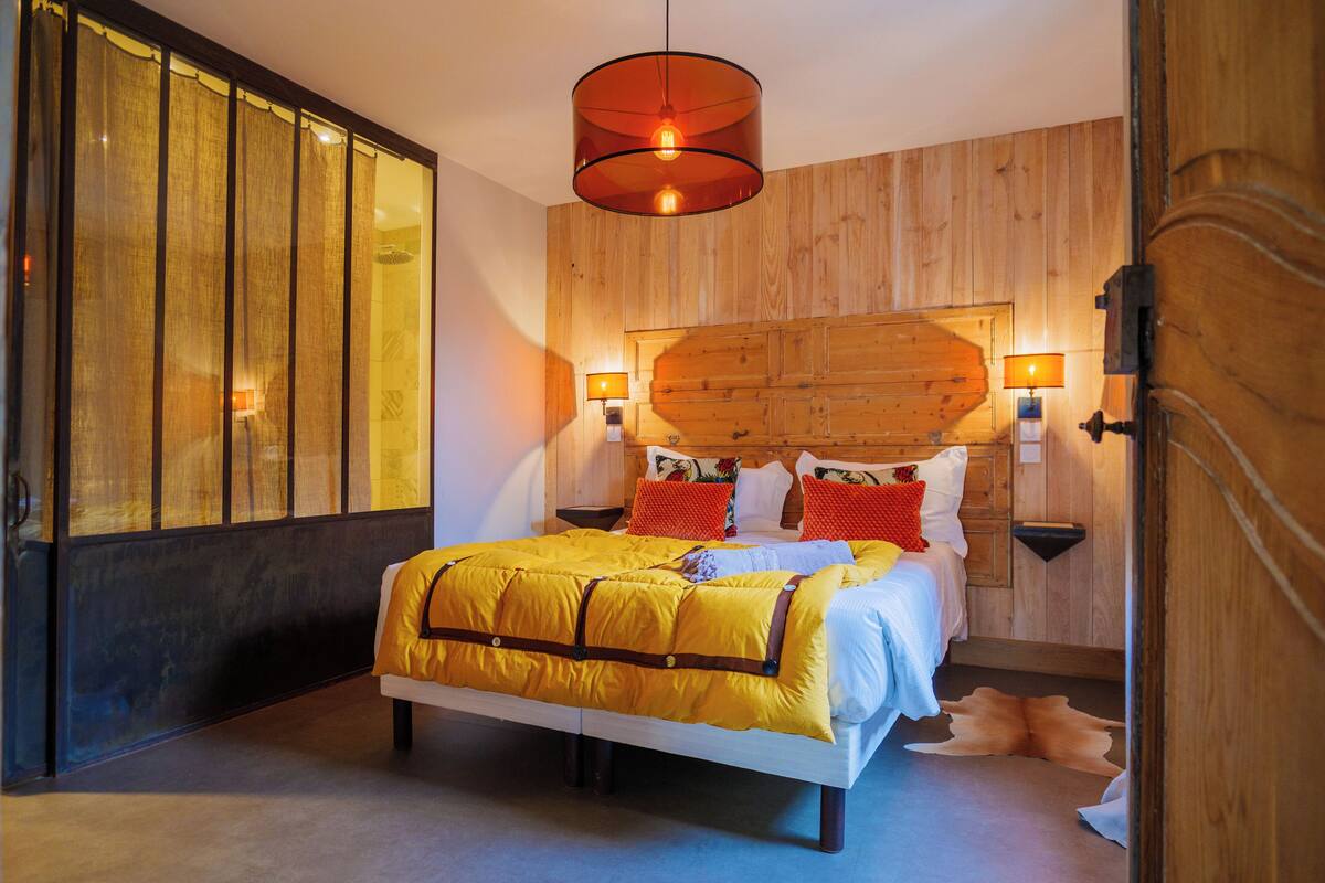 Un dormitorio con cabecera de madera, cama con edredón amarillo, dos lámparas de noche y un ventanal con cortinas.
