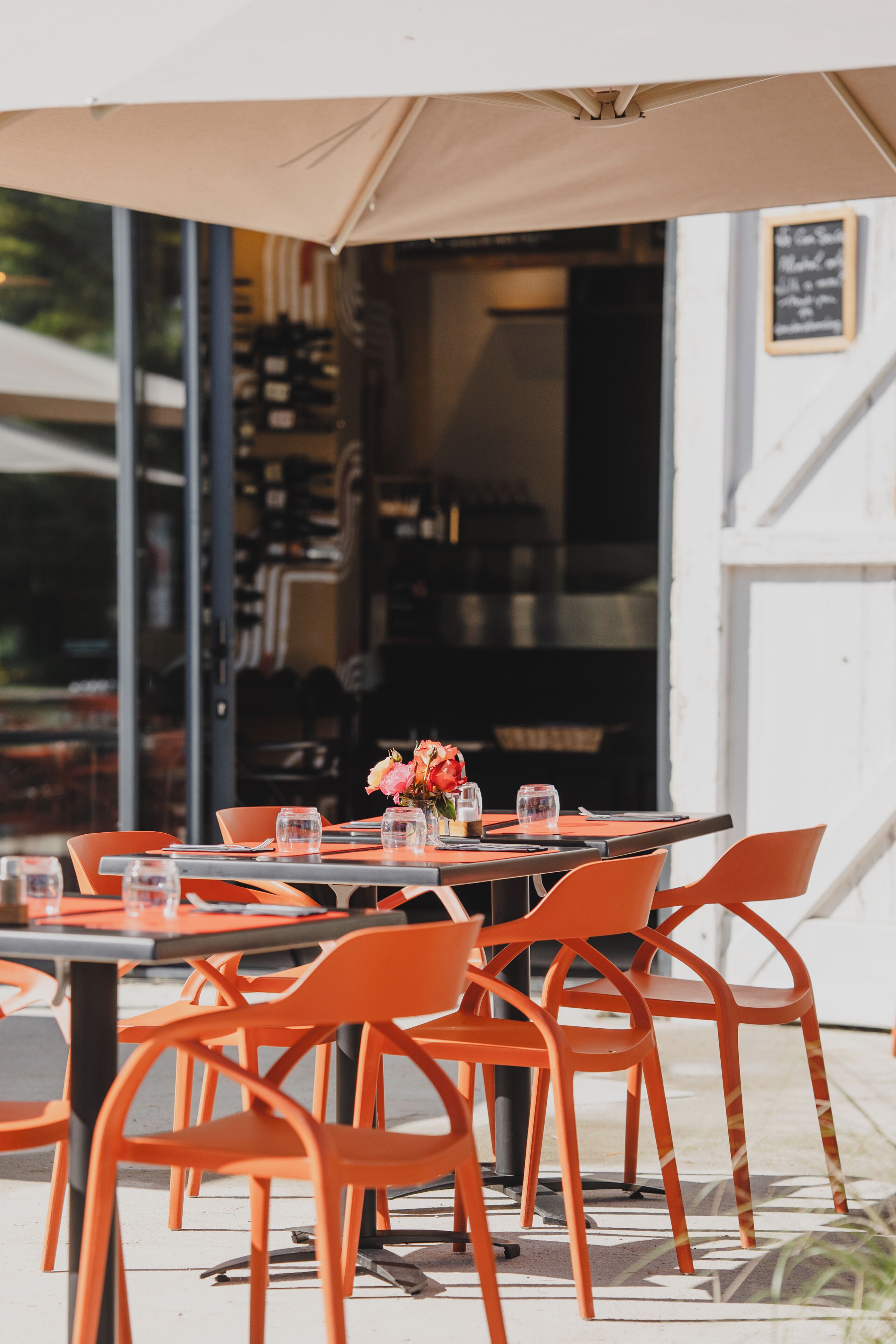 An outdoor dining area with orange chairs and tables, a large umbrella, and a view of the interior through glass doors.