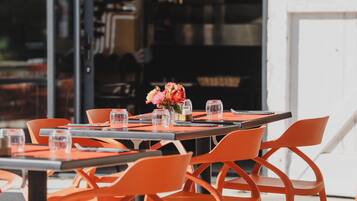 An outdoor dining area with orange chairs and tables, a large umbrella, and a view of the interior through glass doors.