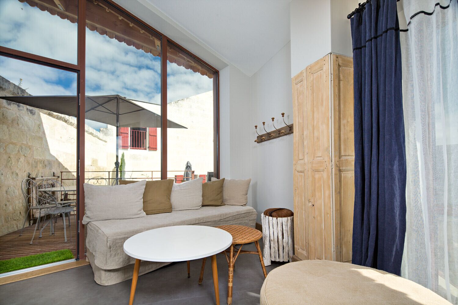 Un salon chaleureux avec un canapé, une table ronde et un tabouret en osier. Une grande fenêtre offre une vue sur un immeuble et un balcon.