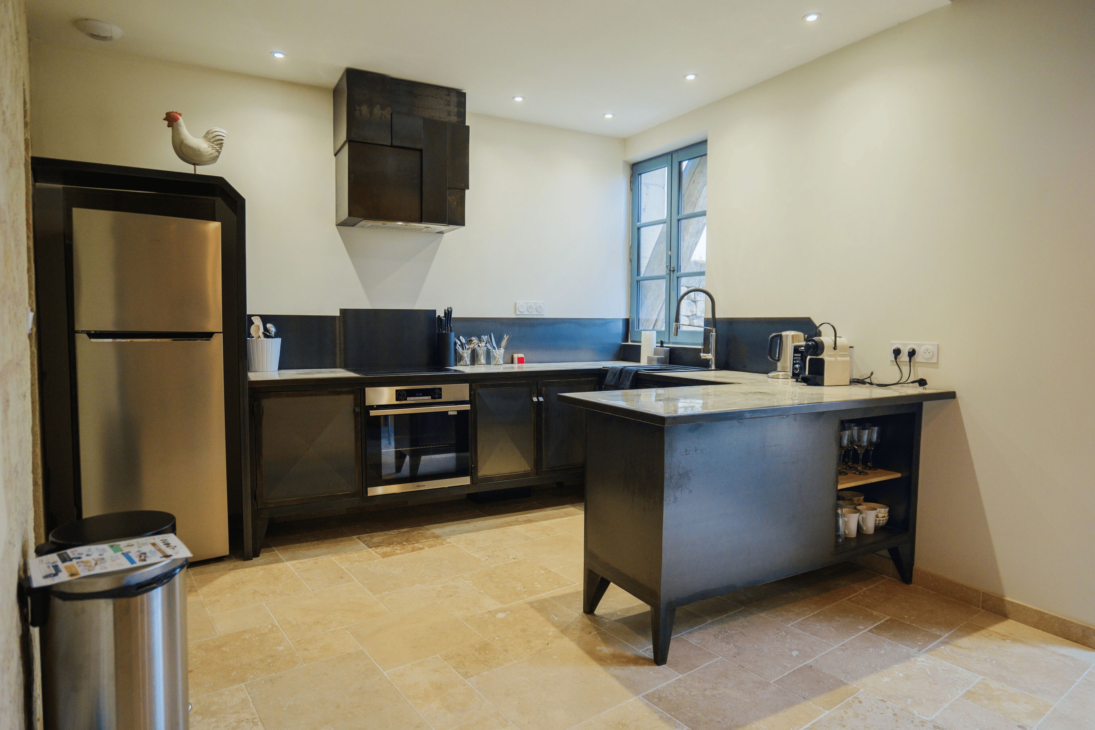 A modern kitchen with a stainless steel refrigerator, oven, and sink, a window with blue shutters, and a rooster decoration on the wall.