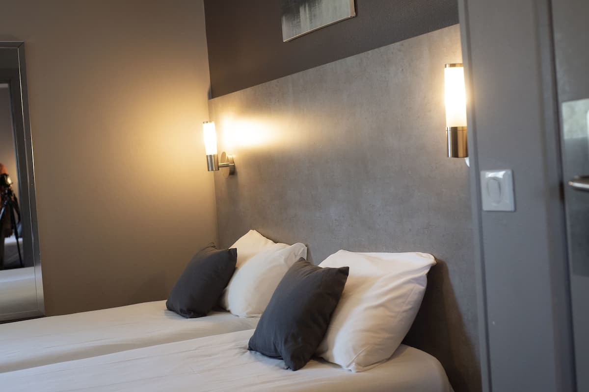 Une chambre d’hôtel avec deux lits, chacun agrémenté d’oreillers blancs et gris foncé, et deux appliques murales.