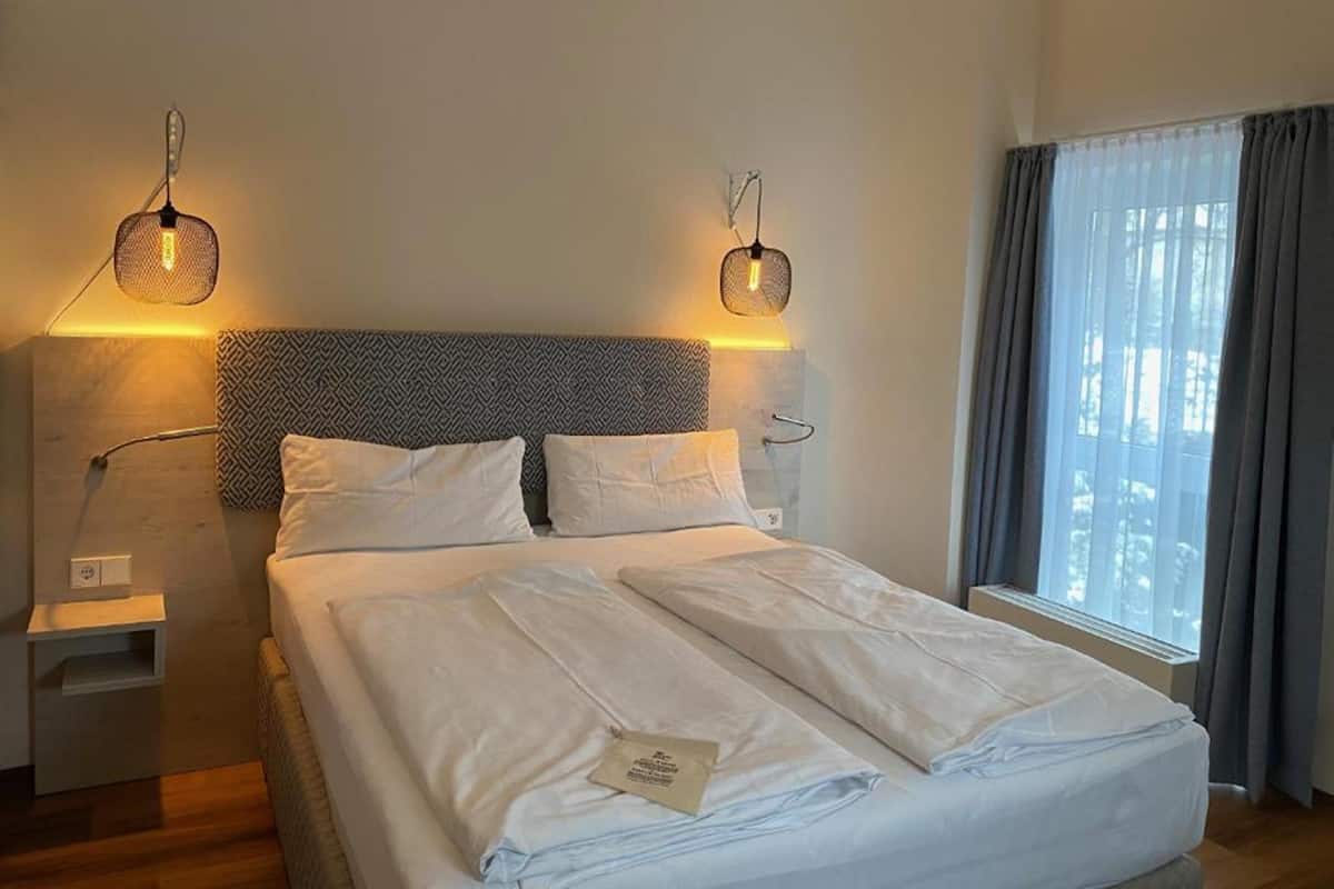 A neatly made bed with white linens, a headboard with a geometric pattern, and two wall-mounted lamps with woven shades.