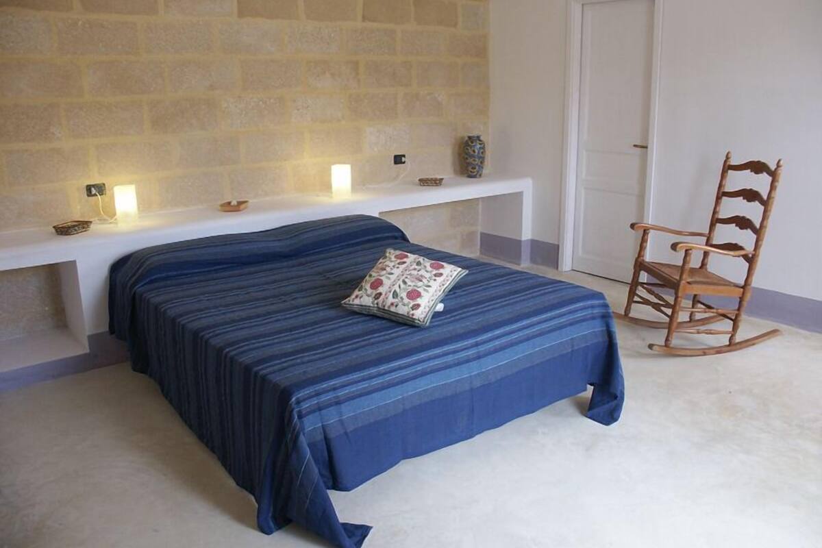 A bedroom with a bed, a bench, a chair, and a wall with a light fixture.