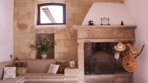 A cozy room with a stone fireplace, a striped sofa, a small table with a vase of flowers, and a collection of hats on a stand. - B&B Masseria 12 Granai (Minervino di Lecce)