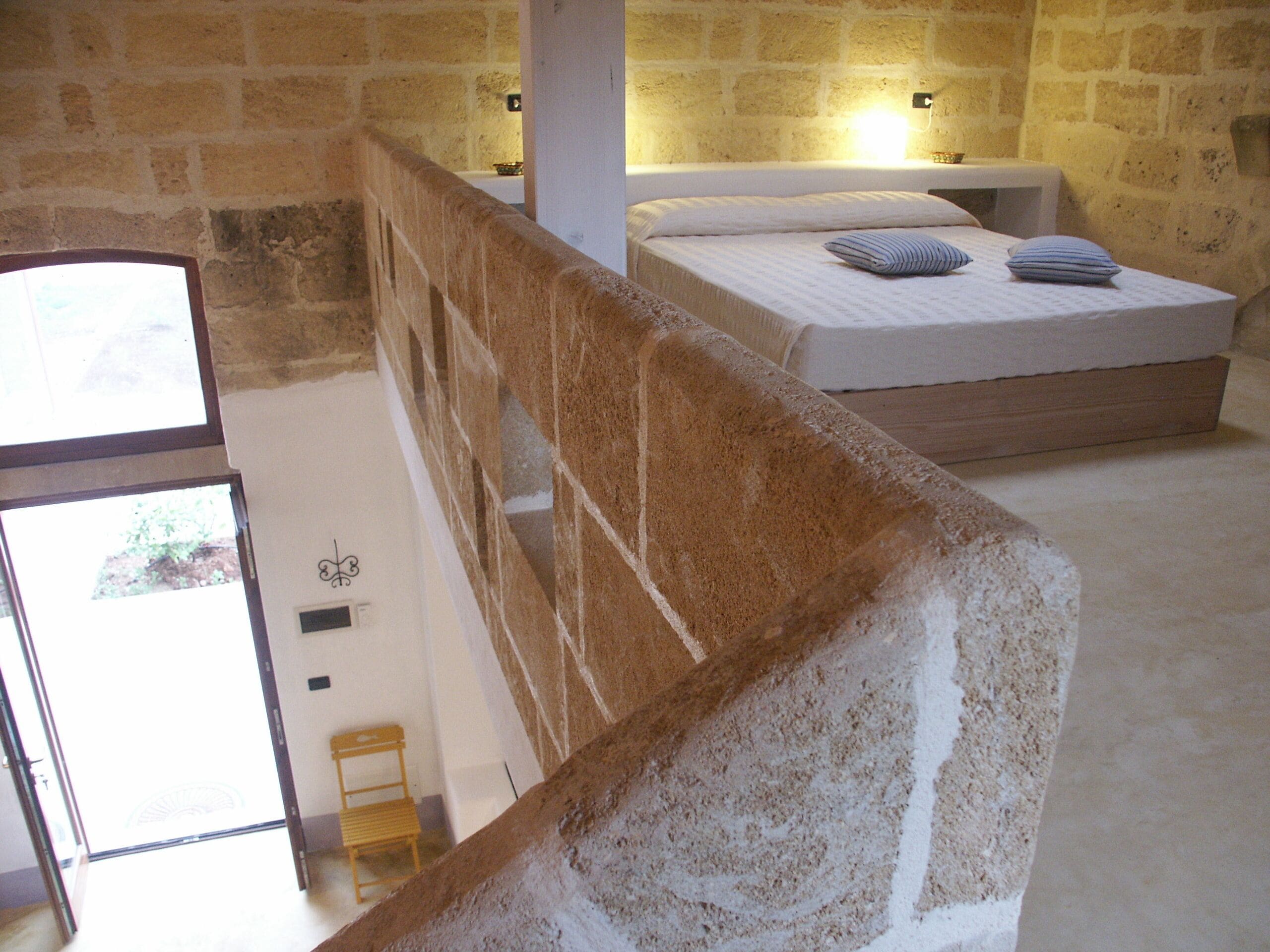 Un dormitorio con pared de piedra, una cama con almohadas y una silla de madera.