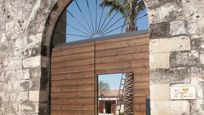 A wooden gate with a stone archway, a sign reading "Masserie dei 12 Granati", and a view of a building with a palm tree in the background. - B&B Masseria 12 Granai (Minervino di Lecce)