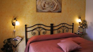 Uma cama com cabeceira, duas luminárias de apoio, um quadro de rosa na parede e uma pequena mesa com um vaso de flores.