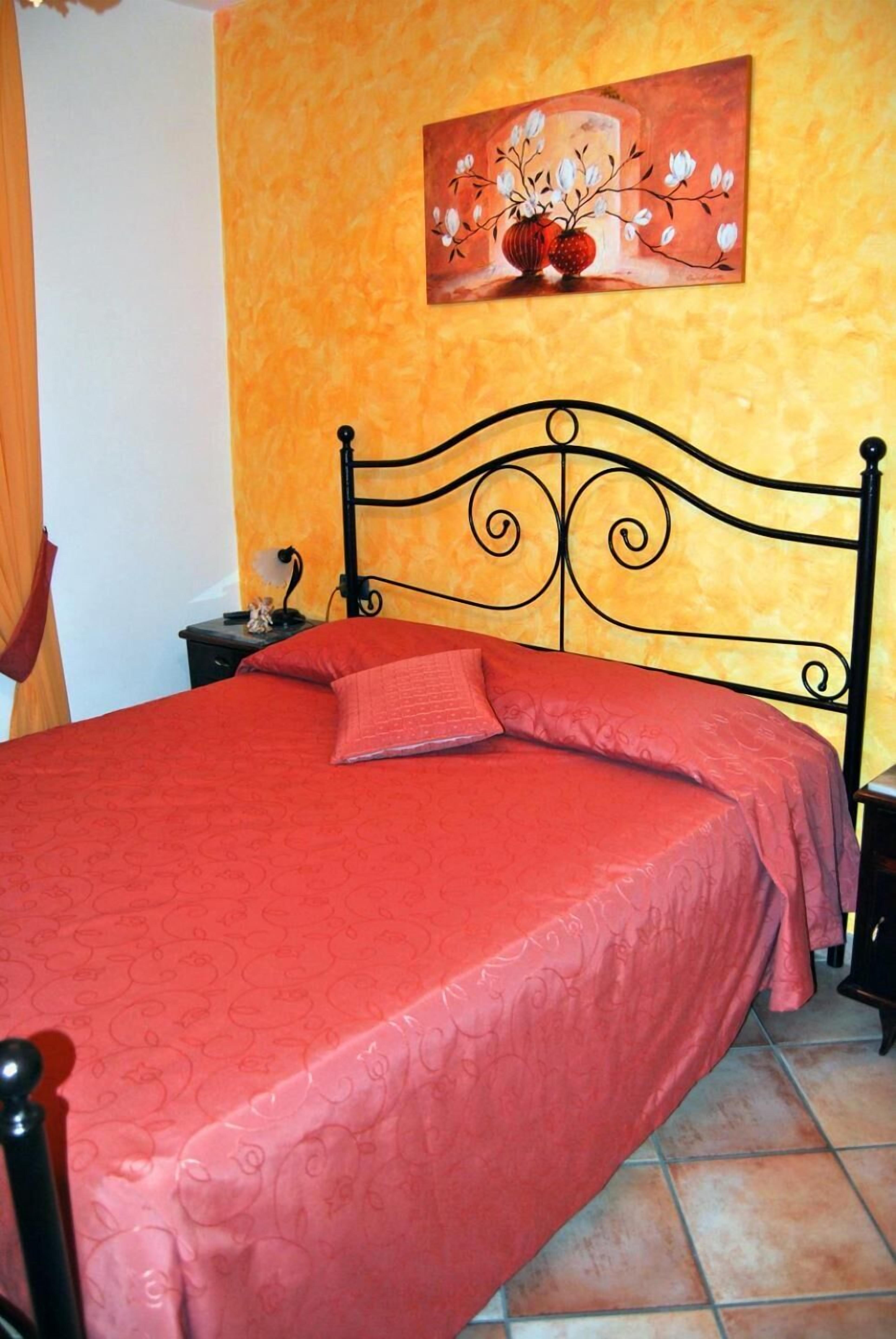 A bed with a red bedspread and pillows, a black metal headboard, a small bedside table with a lamp, and a painting of flowers on the wall.