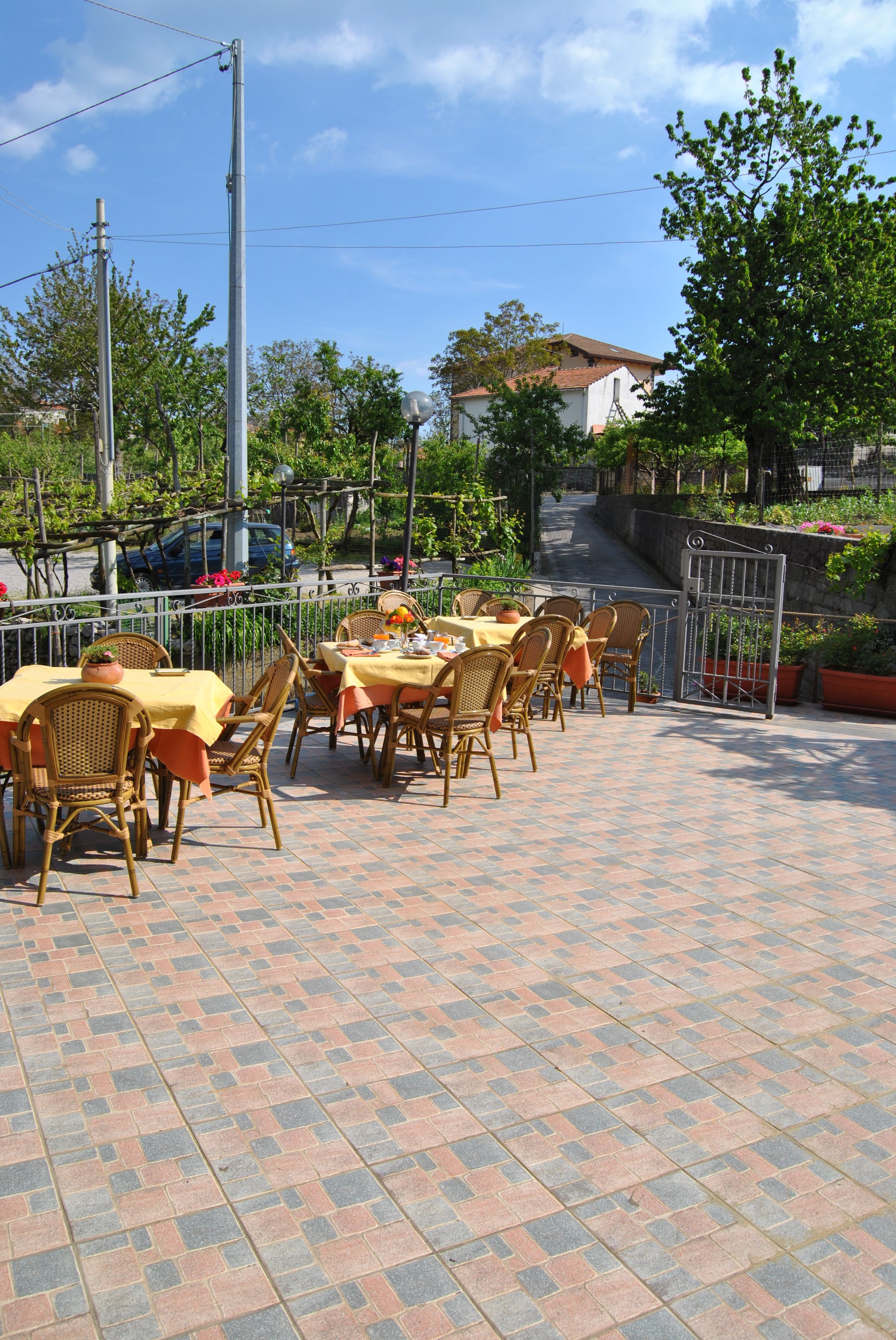 Un café al aire libre con mesas y sillas, piso de ladrillo y vista a una calle y edificios al fondo.