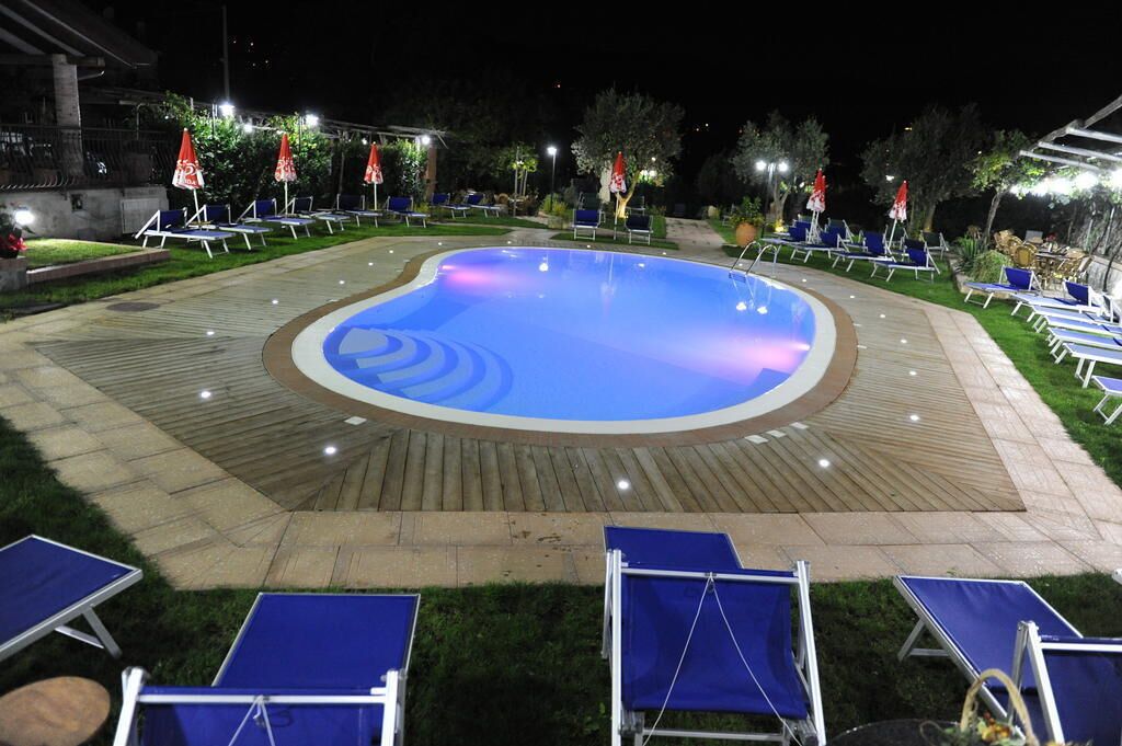A swimming pool surrounded by lounge chairs and umbrellas, with a paved deck and well-lit area.