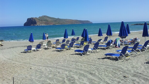 Una playa con filas de sillas y sombrillas azules, un promontorio rocoso al fondo, y mar tranquilo.