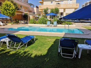 A pool area with lounge chairs and umbrellas, surrounded by a well-maintained lawn and buildings in the background. - Nicolas Studios & Apartments (Chania)