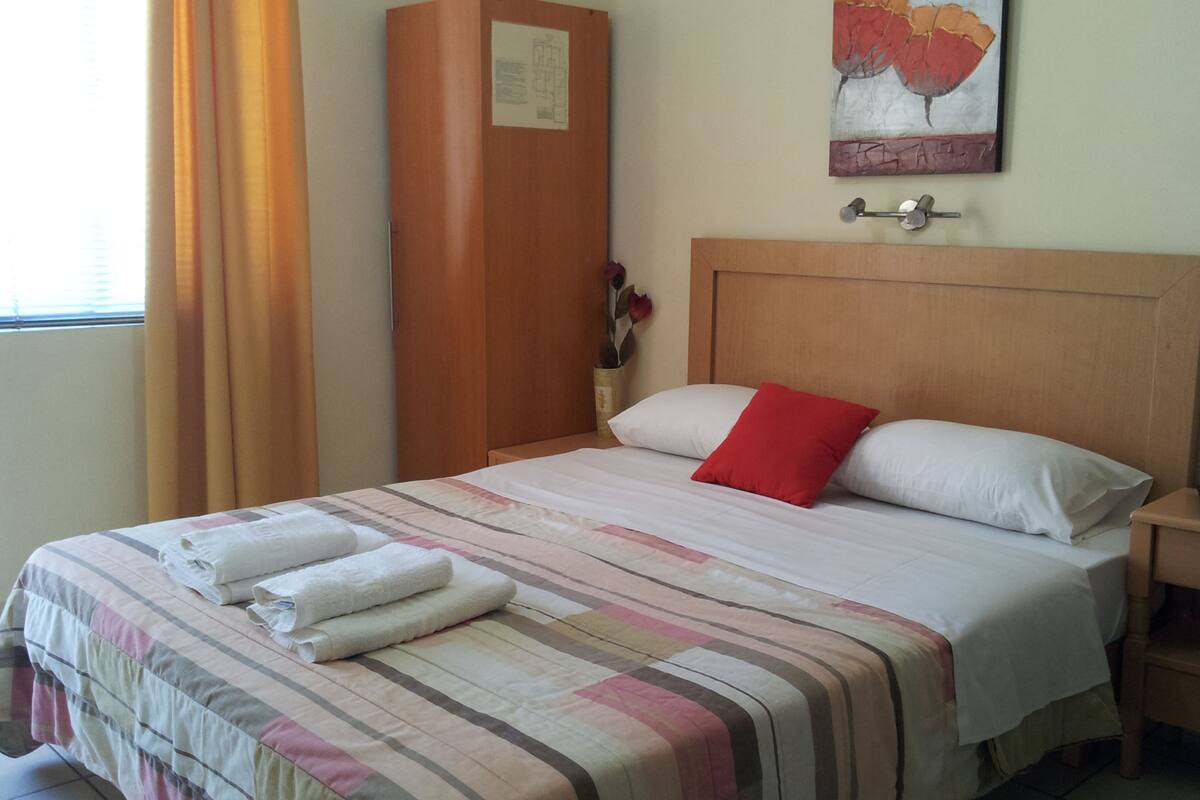 A hotel room with a bed, a nightstand, a wooden wardrobe, a painting, and a window with curtains.