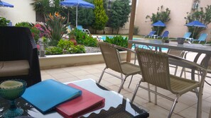 An outdoor seating area with wicker chairs and a table, a pool, and various plants and flowers. - Nicolas Studios & Apartments (Chania)