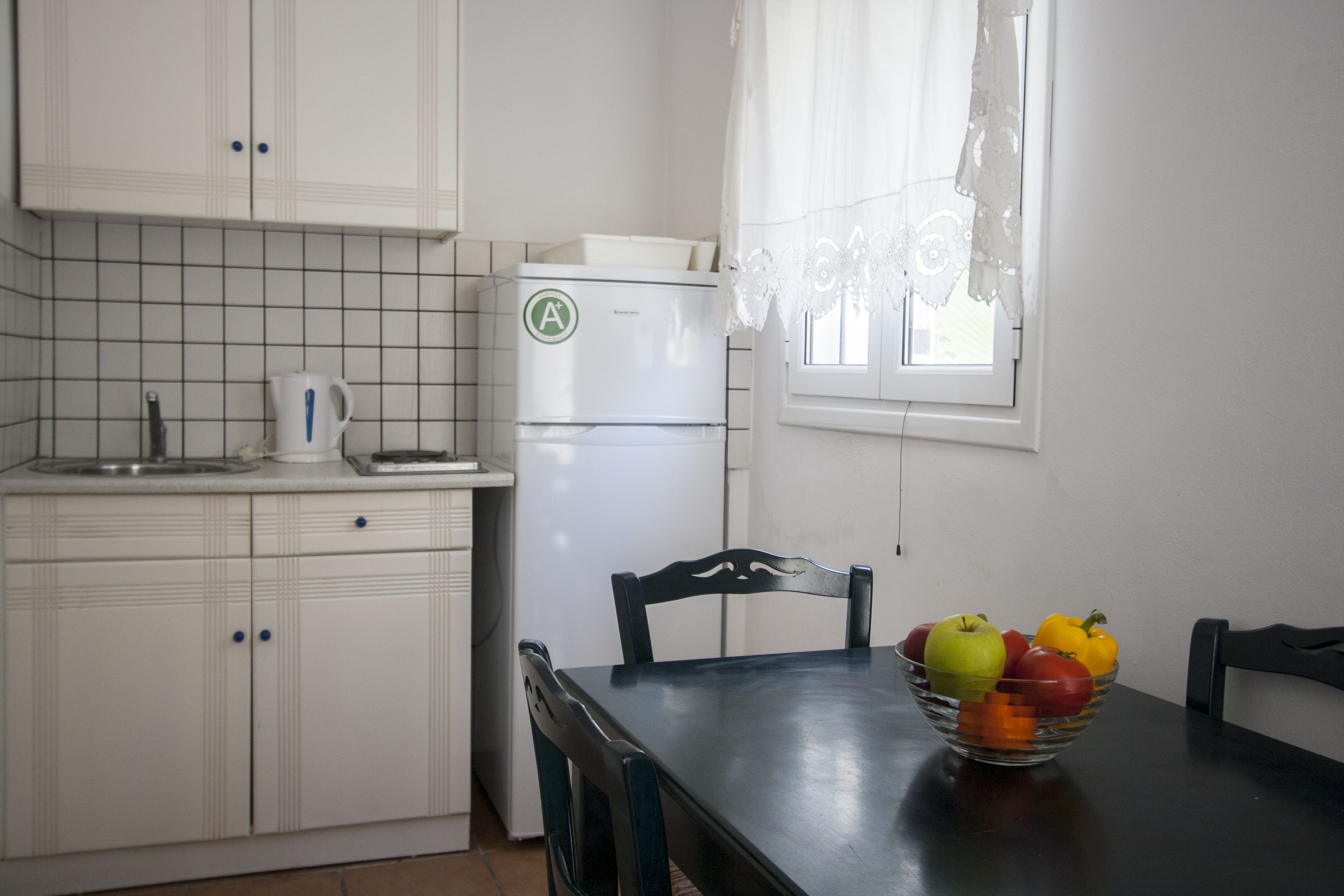 A kitchen with white cabinets, a refrigerator, a kettle, and a bowl of fruit on the table.