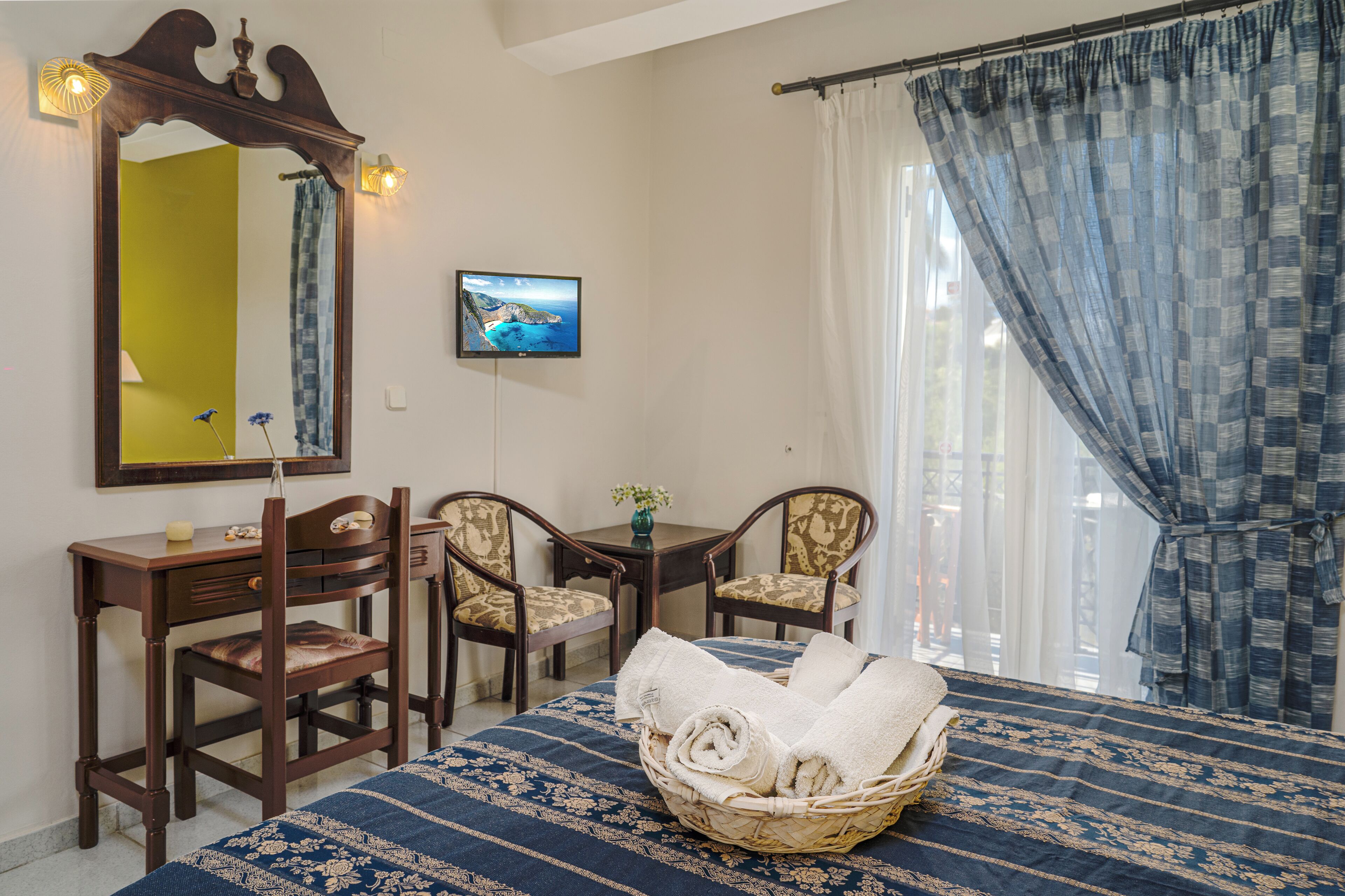 A room with a bed, a desk, two chairs, a mirror, a television, and a window with curtains.