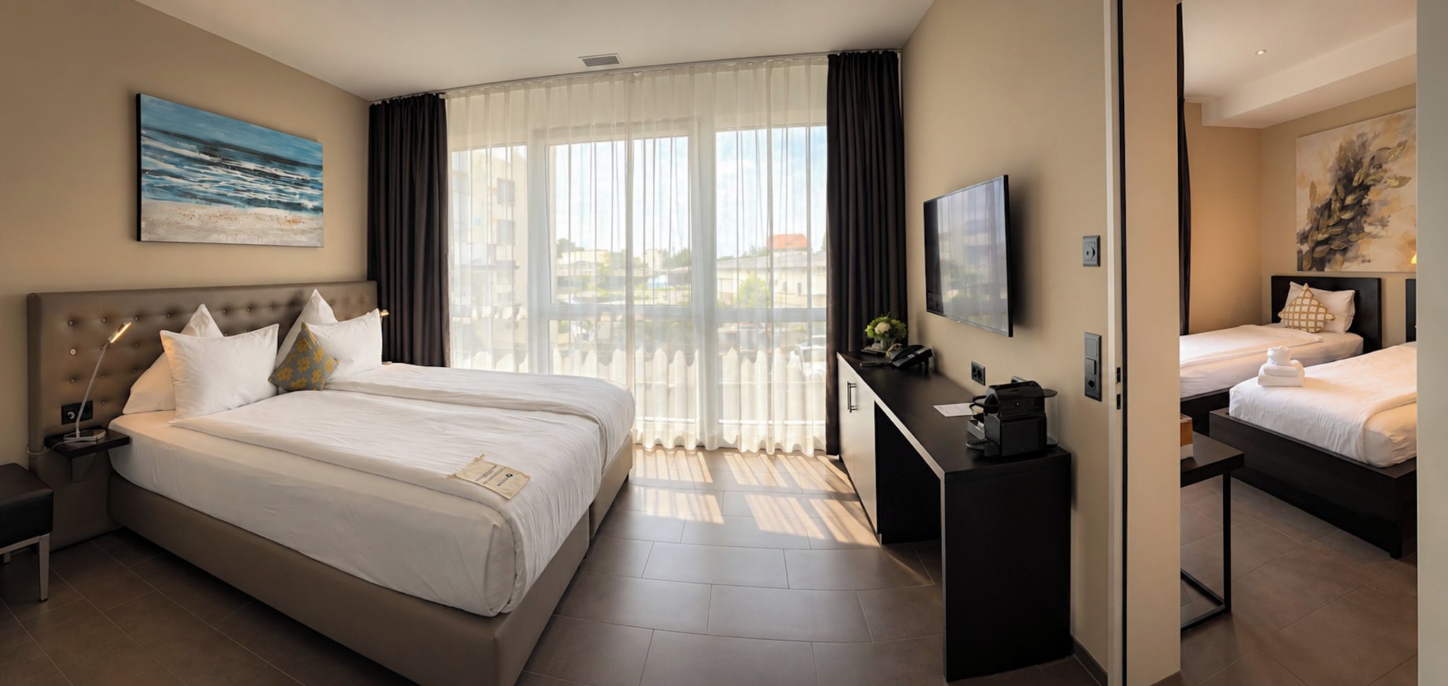 A hotel room with a large bed, a desk with a flat-screen TV, and a view of the city through large windows.