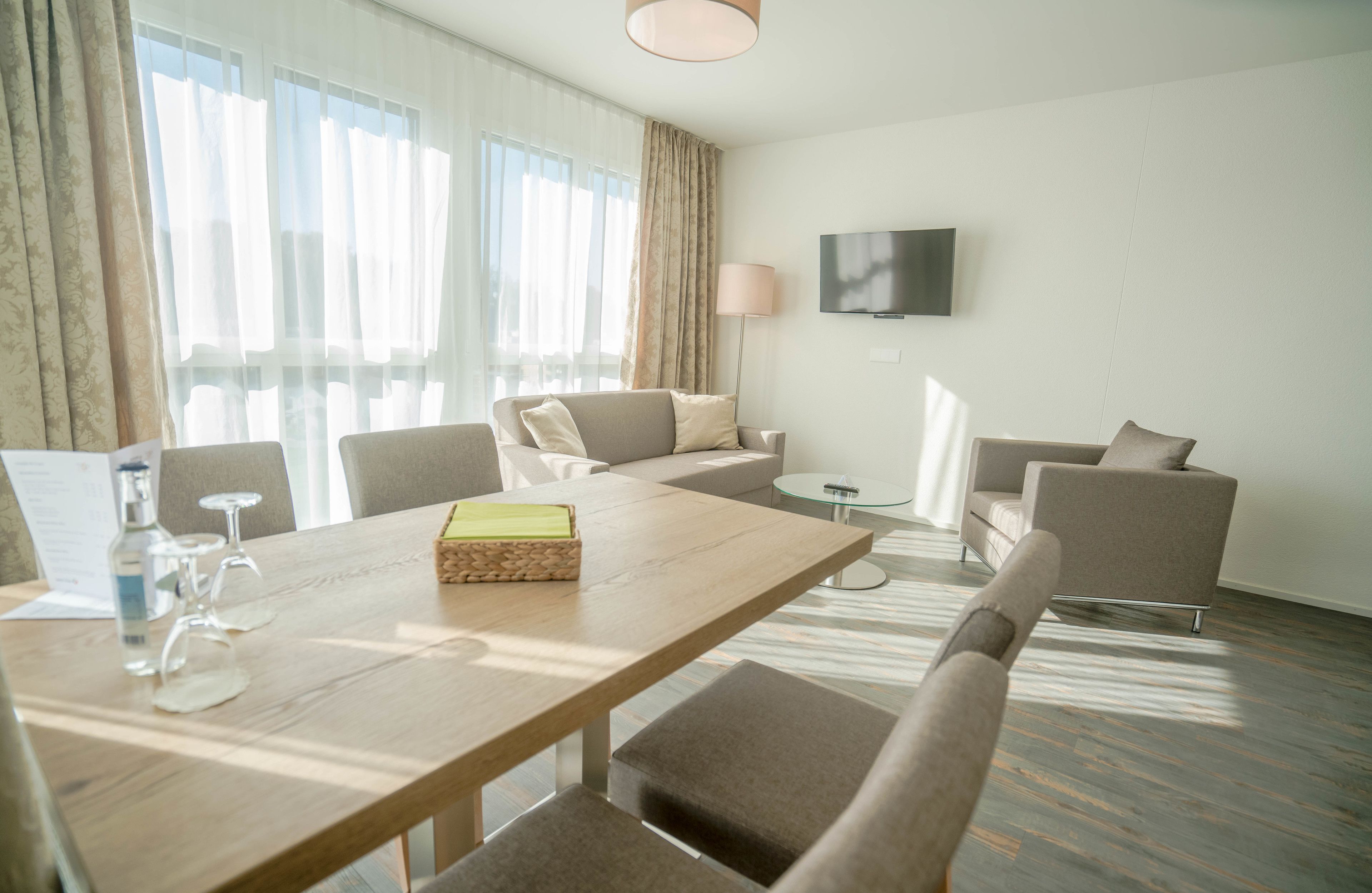 A modern hotel room with a wooden dining table, two sofas, a TV mounted on the wall, and a large window with sheer curtains.