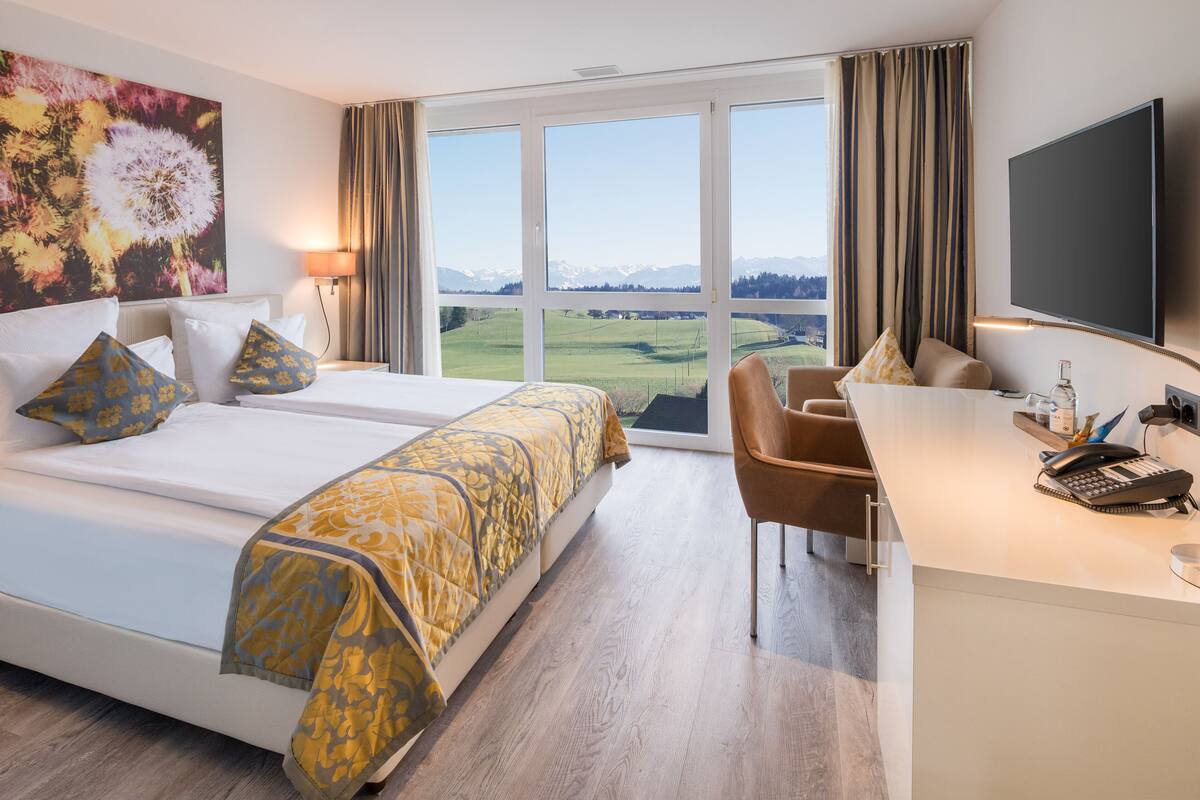 A hotel room with a large bed, a desk with a phone and a lamp, and a view of a green landscape through the window.