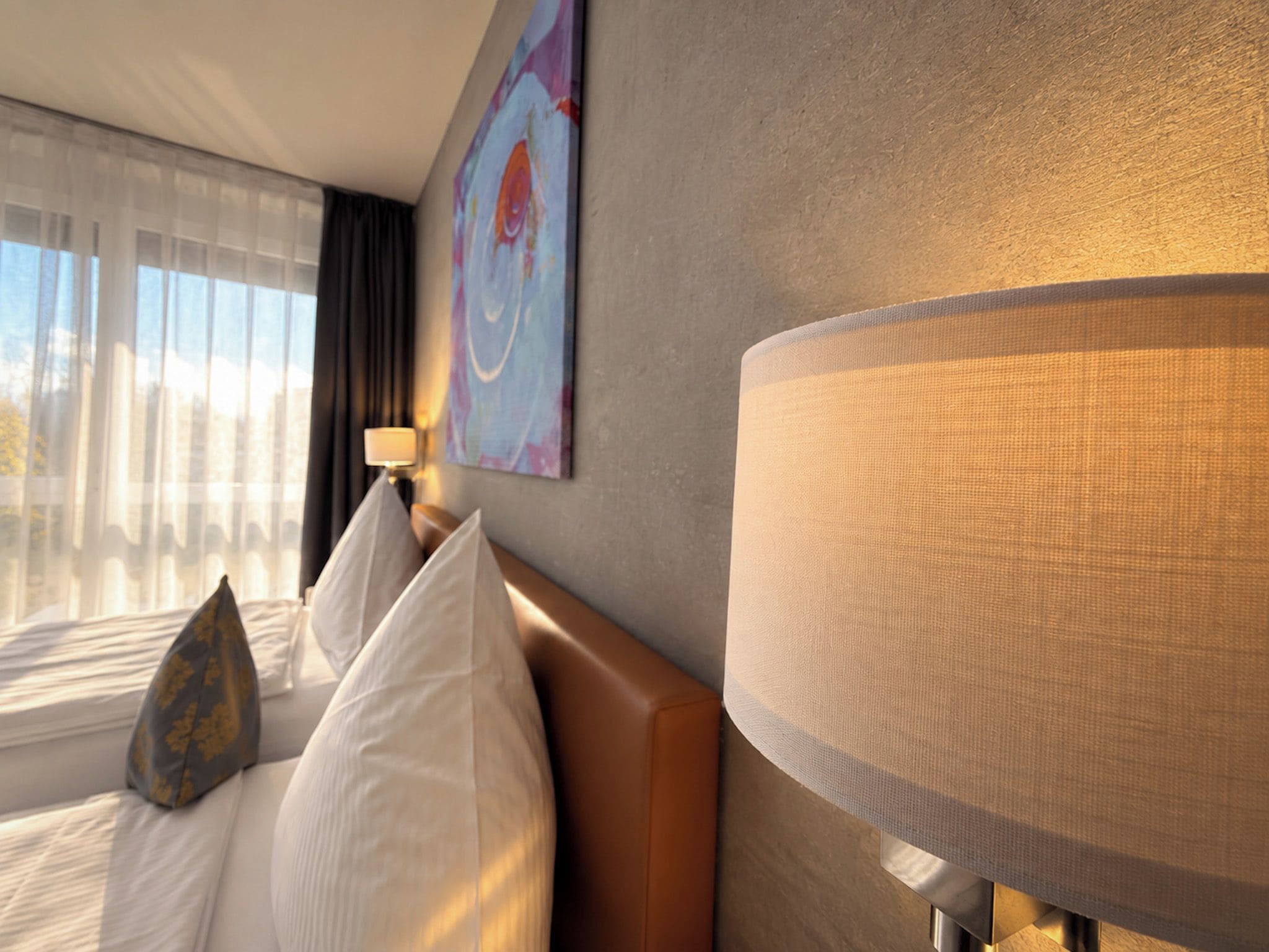 Une chambre d’hôtel avec un grand lit, deux oreillers, une tête de lit, une lampe fixée au mur et une fenêtre avec des rideaux.