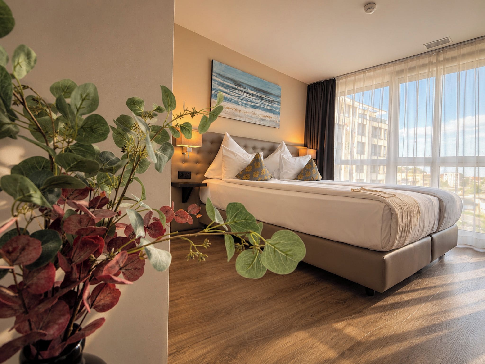 Una habitación de hotel con cama, mesita de noche, lámpara, ventana con cortinas y una planta.