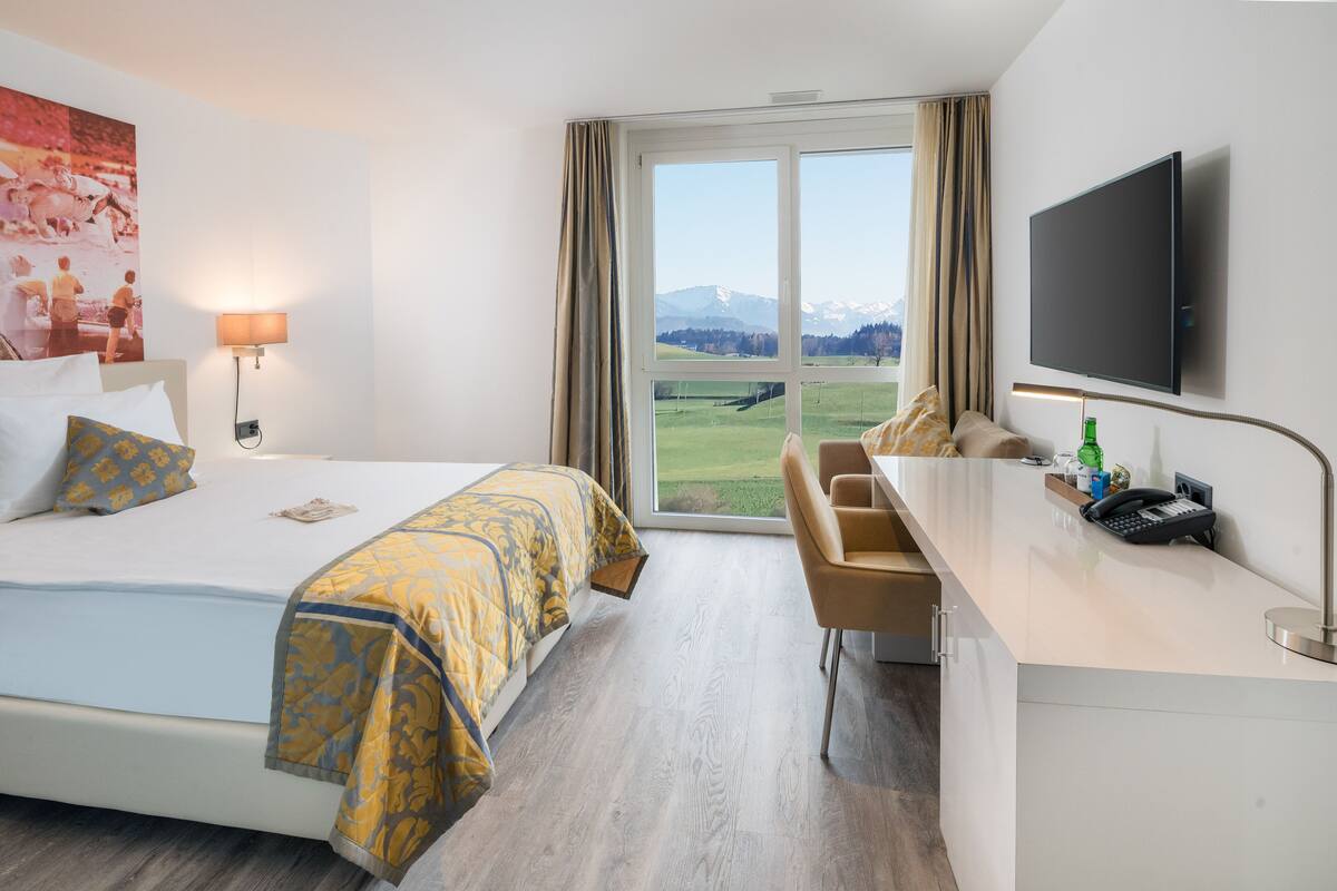 A hotel room with a bed, a desk, a chair, a TV, and a window with a view of mountains.