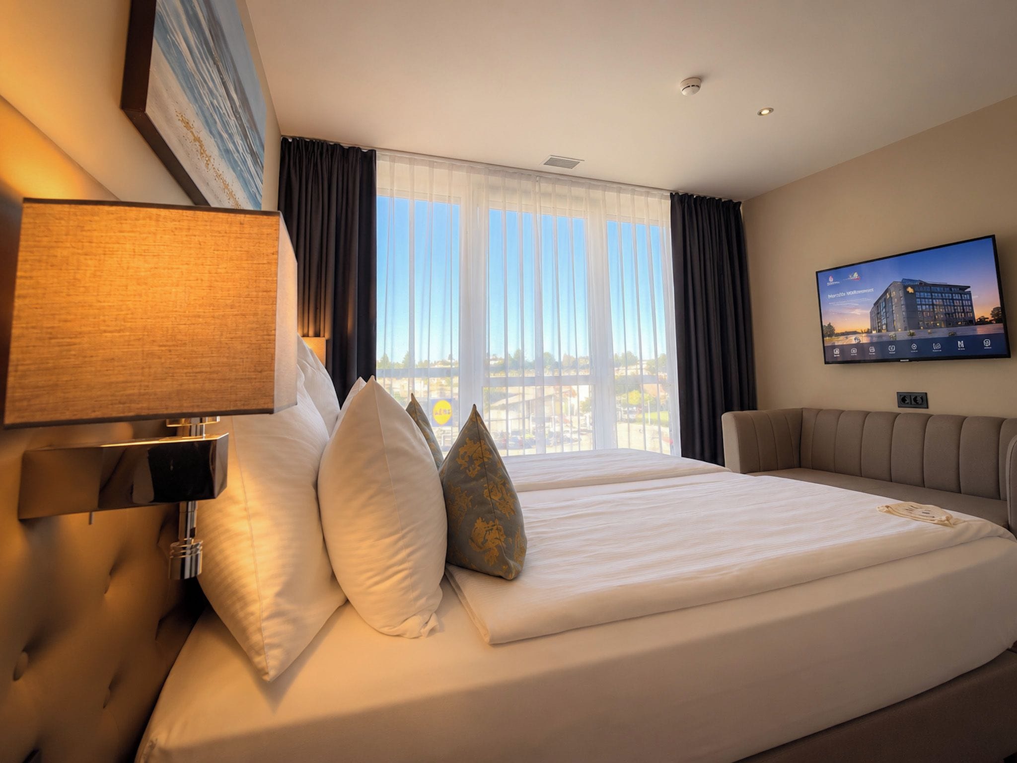 Ein Hotelzimmer mit einem großen Bett, einem Flachbildfernseher, einem Fenster mit Vorhängen und einer Lampe.