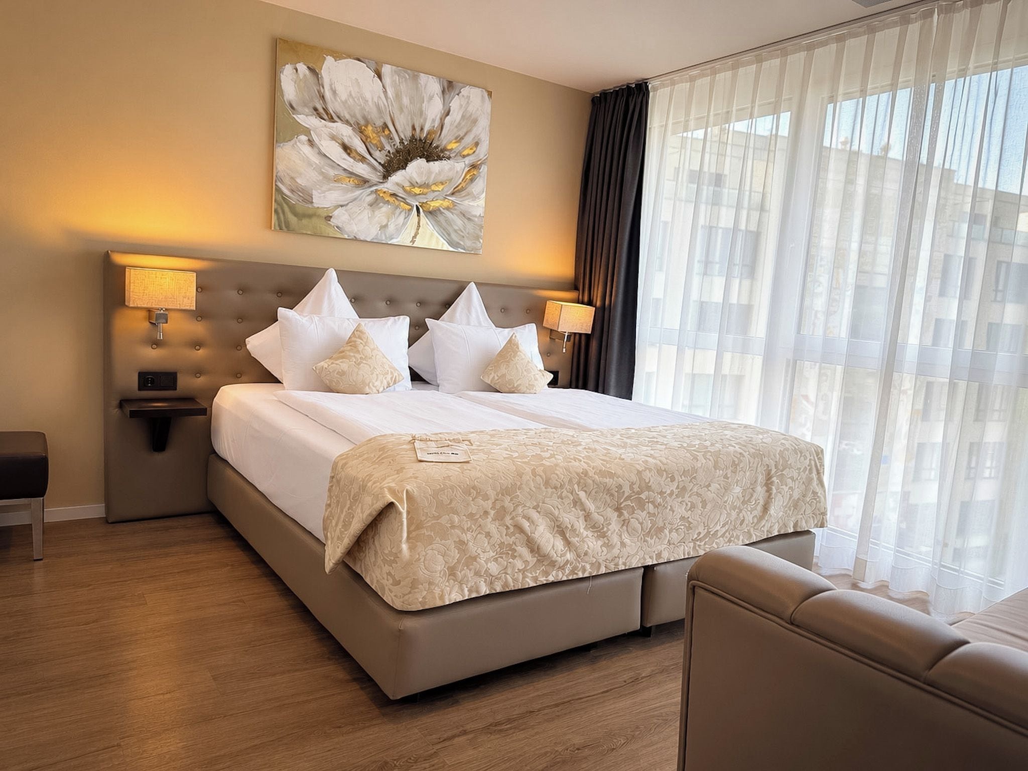 A hotel room with a large bed, two bedside lamps, a headboard, a window with sheer curtains, and a painting above the bed.