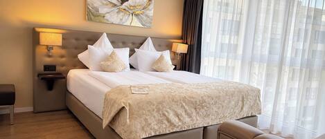 A hotel room with a large bed, two bedside lamps, a headboard, a window with sheer curtains, and a painting above the bed.