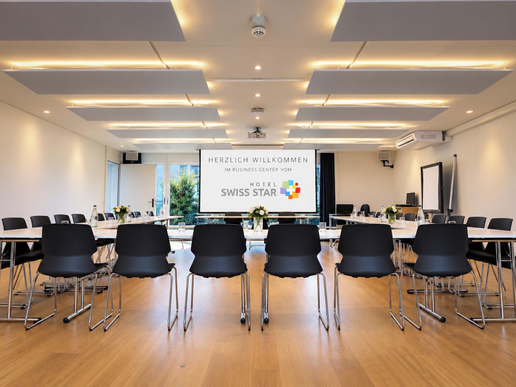 Ein Konferenzraum mit einem langen Tisch, schwarzen Stühlen und einem großen Bildschirm, der eine Willkommensbotschaft zeigt.