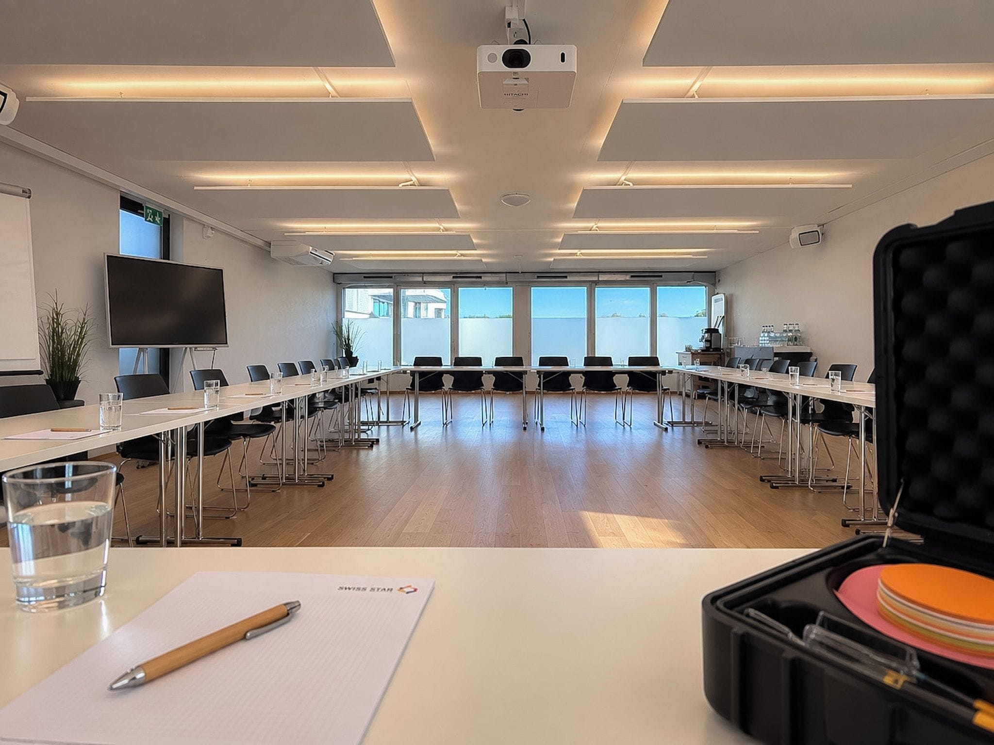 Ein Konferenzraum mit einem langen Tisch, Stühlen, einem Projektor und einem großen Fenster.