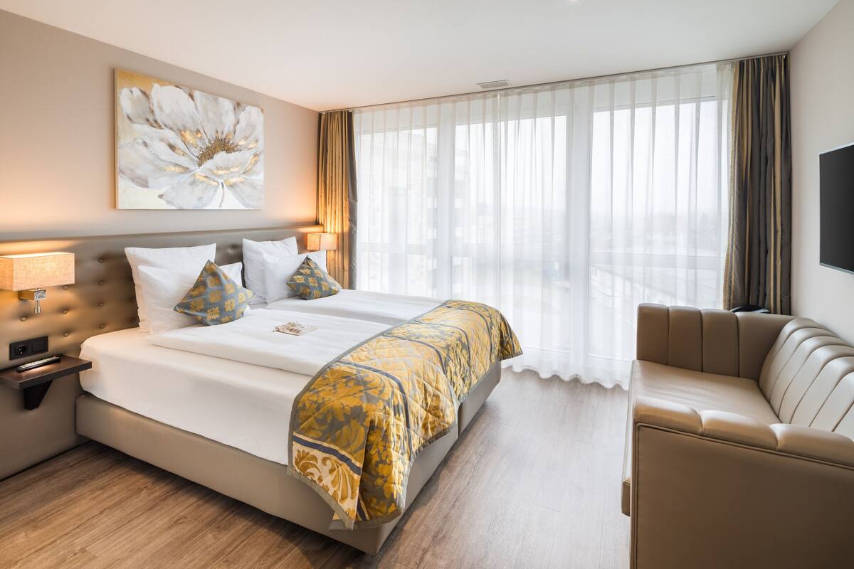 A hotel room with a large bed, a sofa, a flat-screen TV, and a window with sheer curtains.