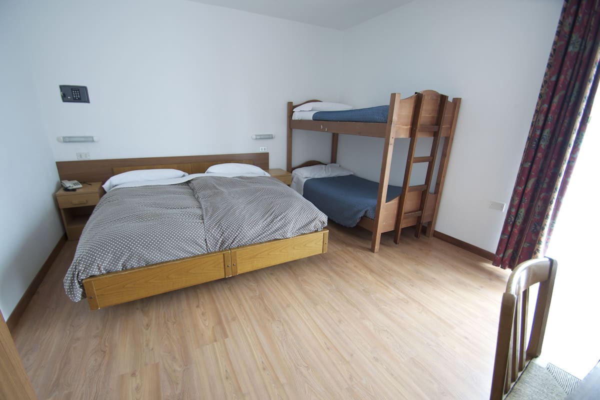 A single bed with a wooden frame and a grey polka dot bedspread, a bunk bed with a blue mattress, a bedside table with a phone, and a chair in a room with wooden flooring and white walls.