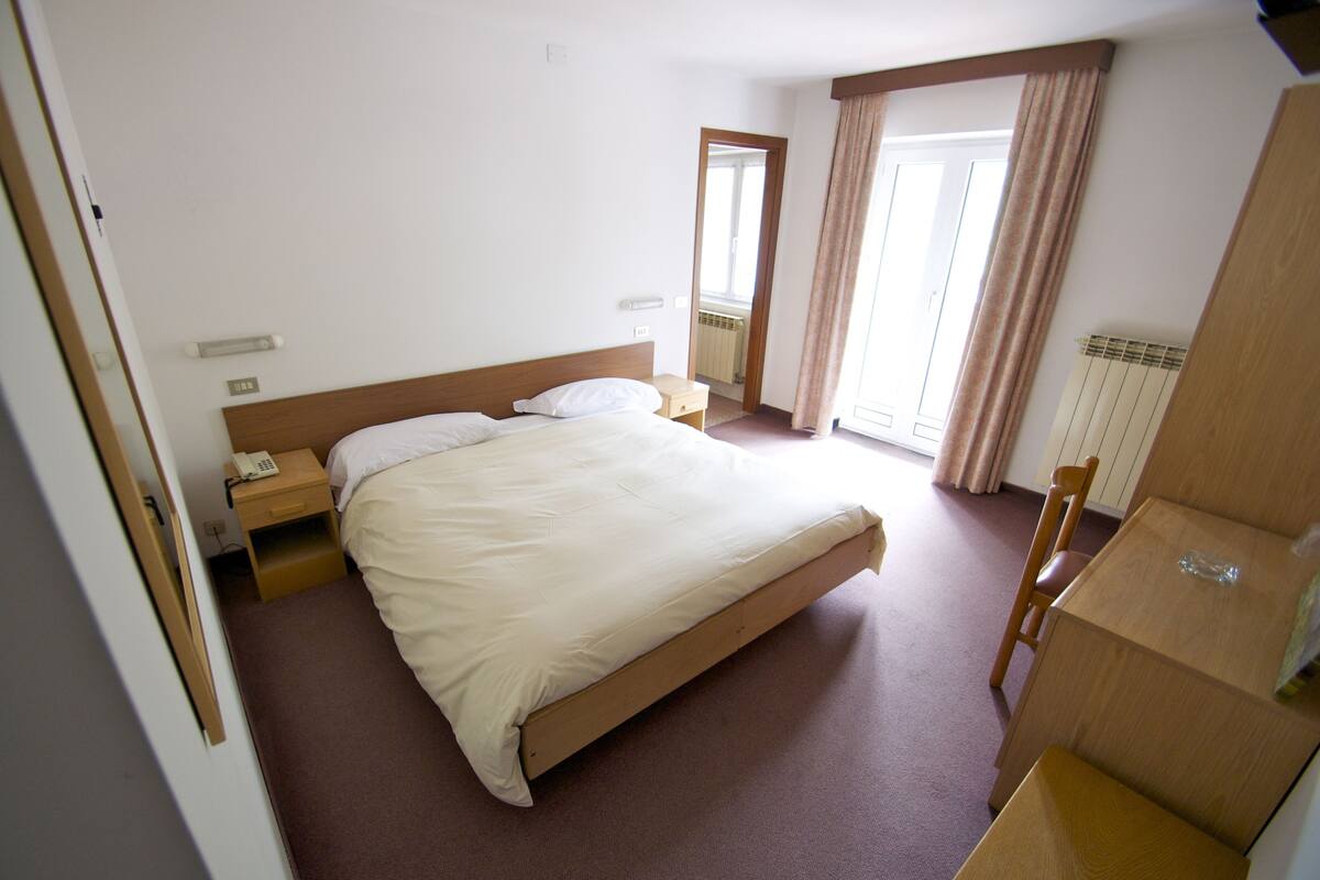 A hotel room with a single bed, a bedside table with a phone, a wooden chair, and a radiator.