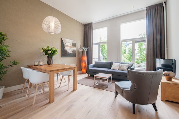Un salon moderne avec une table à manger en bois, des chaises blanches, un canapé gris et un fauteuil marron. On y trouve une grande fenêtre avec des rideaux, un lampadaire et un vase rempli de fleurs.