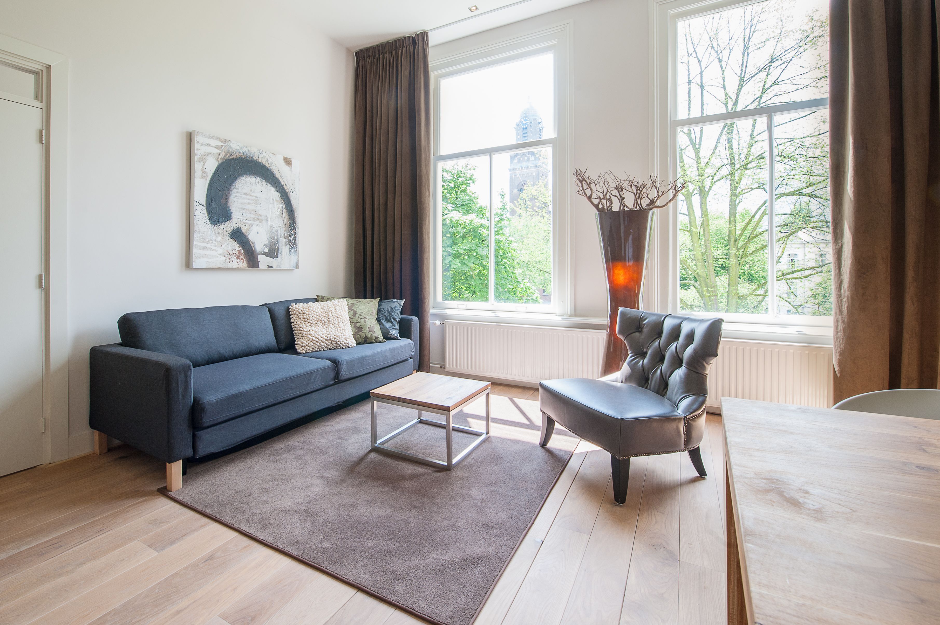 A modern living room with a dark blue sofa, a wooden coffee table, a leather armchair, and a large window with a view of trees and buildings.