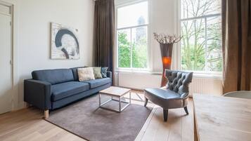 A modern living room with a dark blue sofa, a wooden coffee table, a leather armchair, and a large window with a view of trees and buildings.