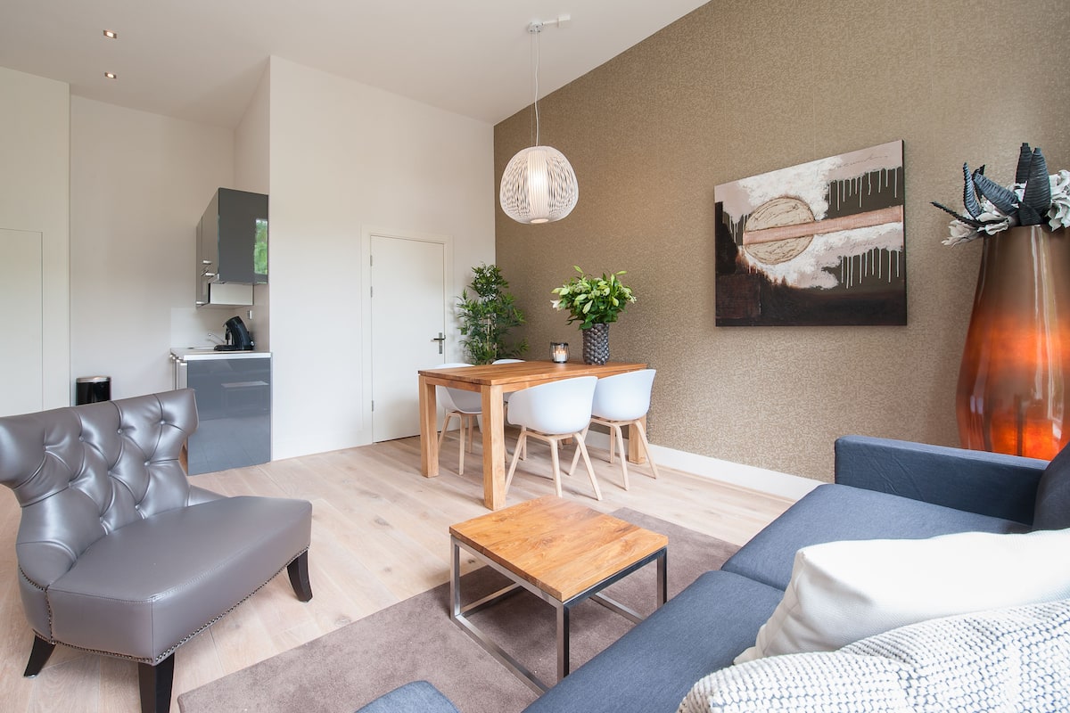 Una sala de estar moderna con un sofá gris, una mesa de centro de madera y un sofá azul. Hay un comedor con una mesa de madera y sillas blancas. Un cuadro grande adorna la pared, y una planta en maceta está sobre la mesa.