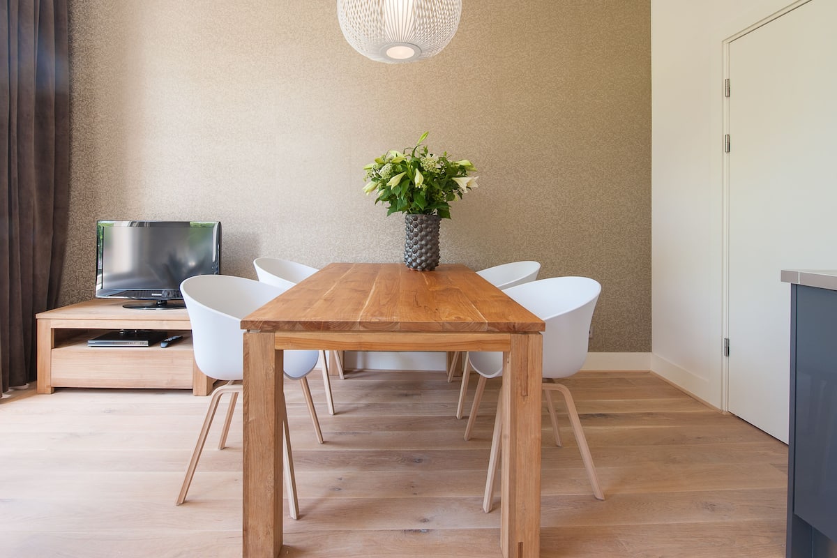 Un comedor con una mesa de madera, sillas blancas, un jarrón con flores, un televisor sobre un mueble bajo y un armario.