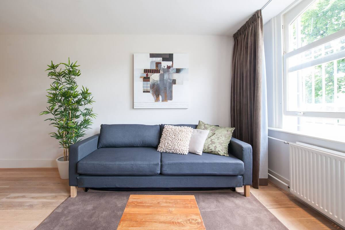 Una sala de estar moderna con un sofá azul, una mesa de centro de madera, una planta en maceta y un ventanal con cortinas.
