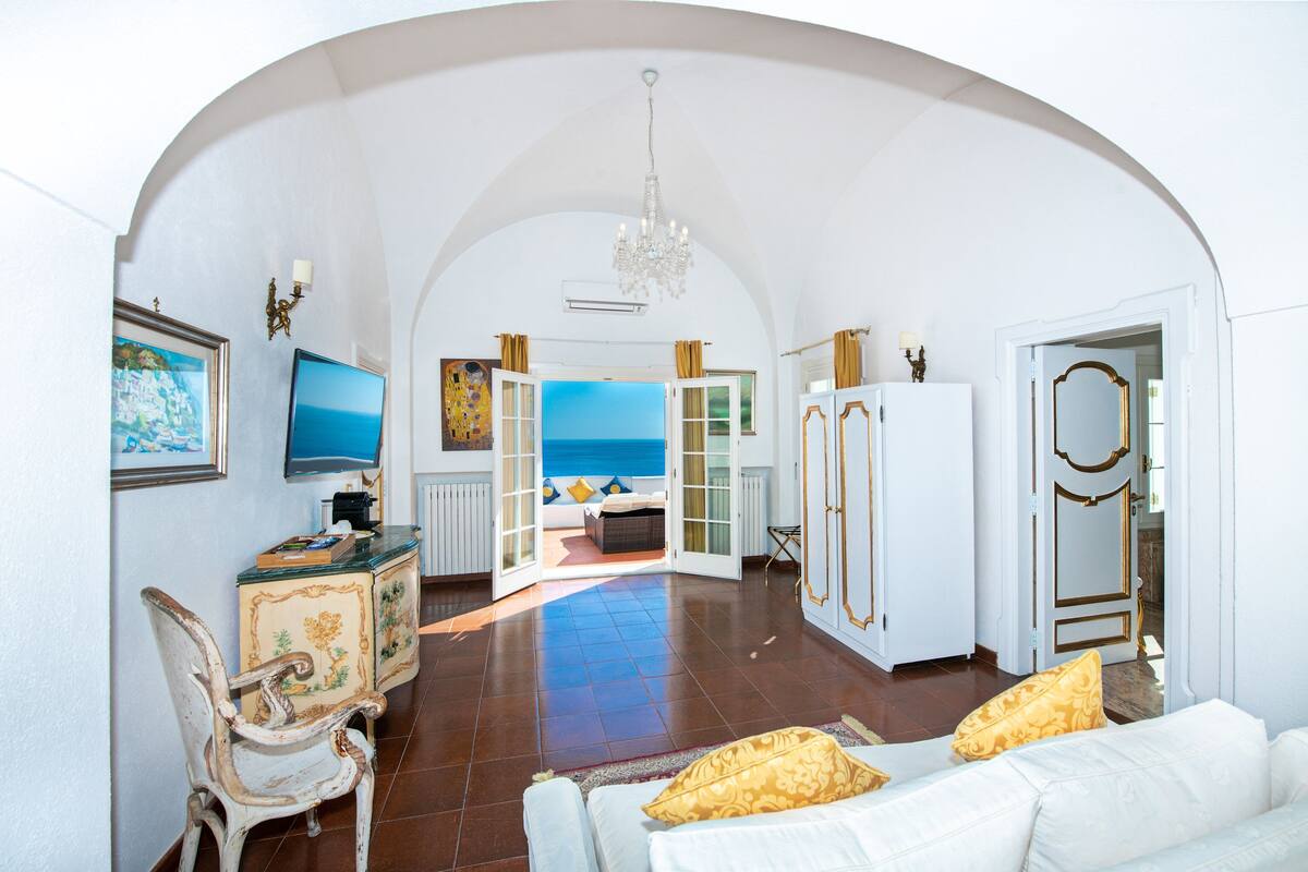 A spacious living area with a view of the sea, a chandelier, and a white sofa with yellow pillows.