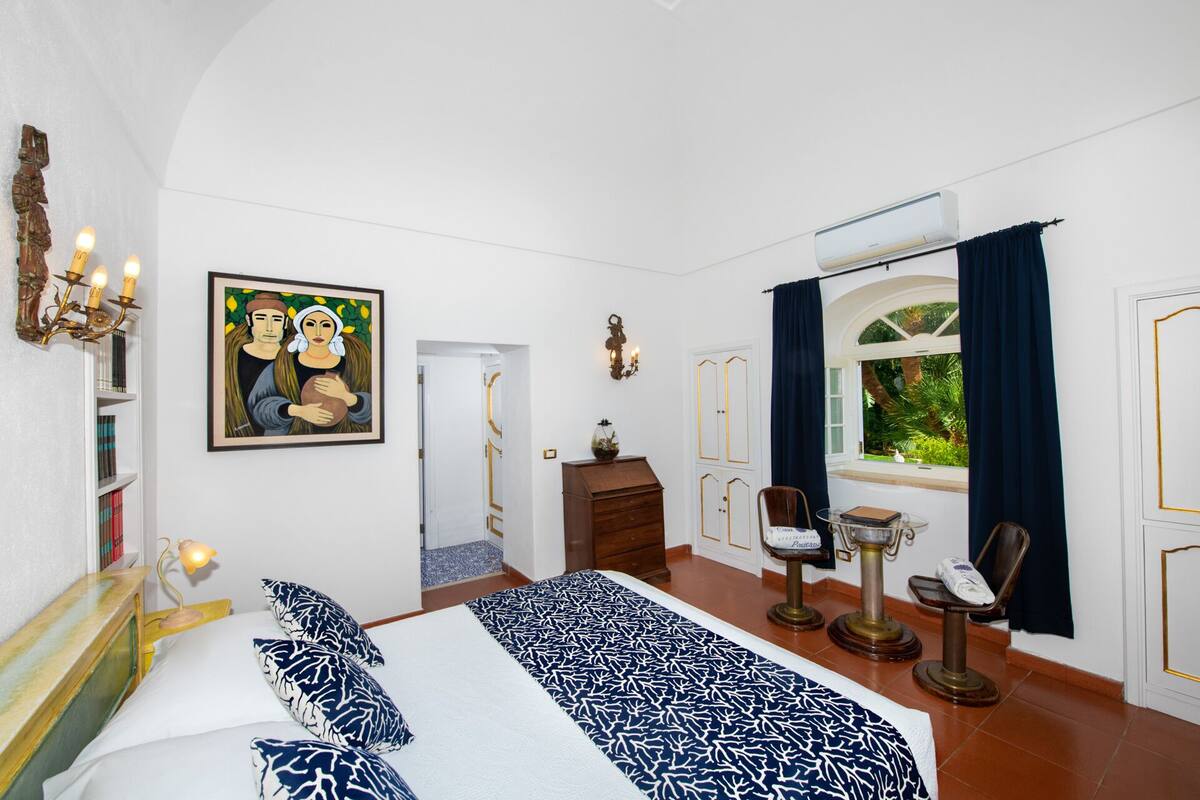 A bedroom with a large bed, a desk with a chair, a window with curtains, a wall-mounted air conditioner, and a painting.