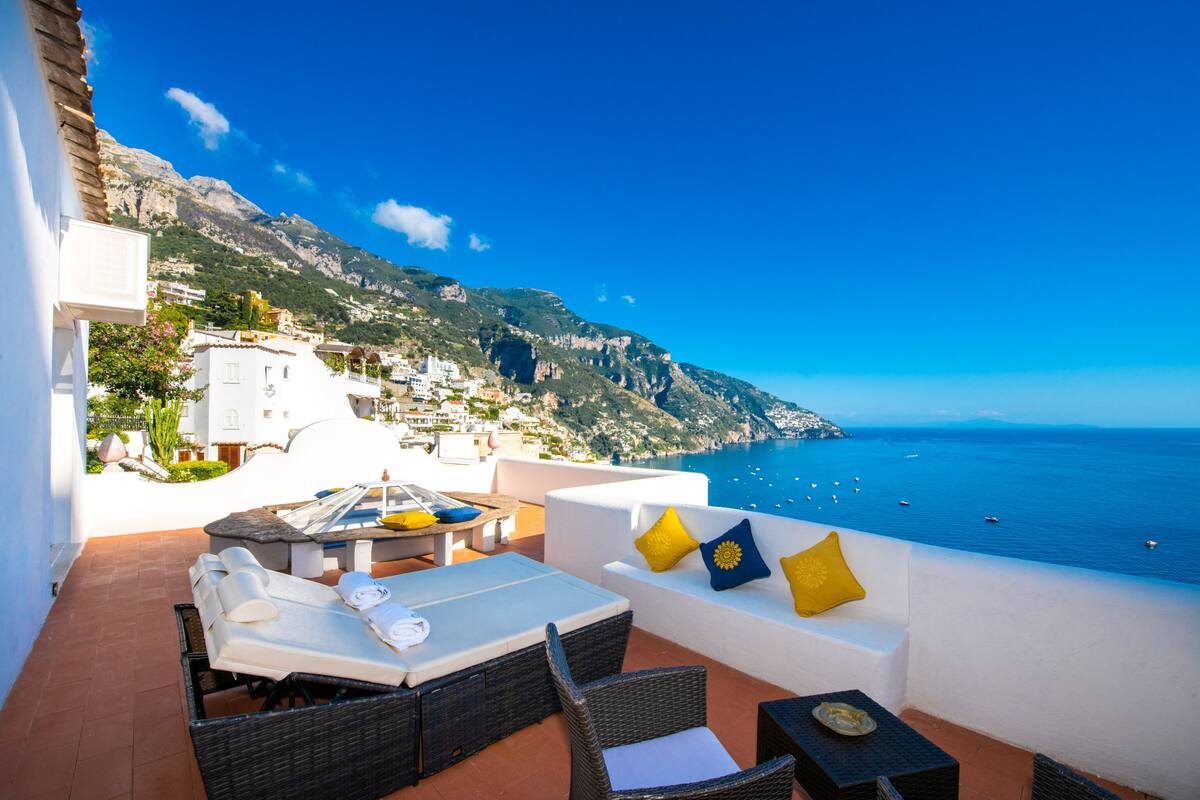 A terrace with outdoor seating, a pool, and a view of the sea and mountains.