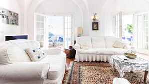 Un salon spacieux, meublé de blanc, avec un tapis à motifs et une vue dégagée sur l’extérieur grâce à une porte ouverte.