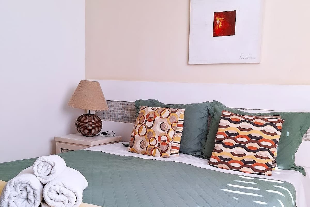 A neatly made bed with green bedding and multiple patterned pillows, a bedside lamp, and rolled towels on the nightstand.