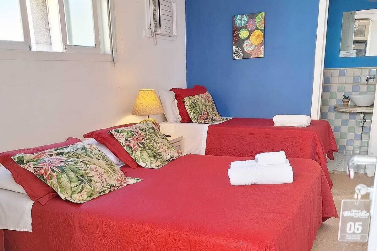 A room with two beds, each with a red bedspread and floral pillows, a blue wall, a small painting, and a bathroom visible through an open door.