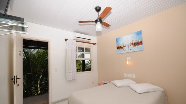 A bedroom with a bed, a ceiling fan, a window with a view of greenery, and a painting on the wall. - Pousada Areias Brancas (Búzios)