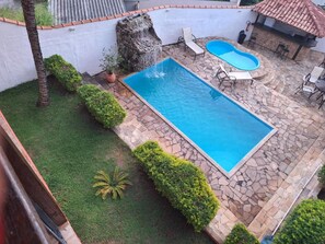 A rectangular swimming pool with a stone border, surrounded by a grassy area and a few small bushes. - Pousada Areias Brancas (Búzios)