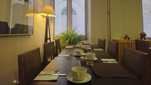 Un comedor con una mesa dispuesta para cuatro personas, sillas, una lámpara, una planta en maceta y una ventana con vista a un edificio.